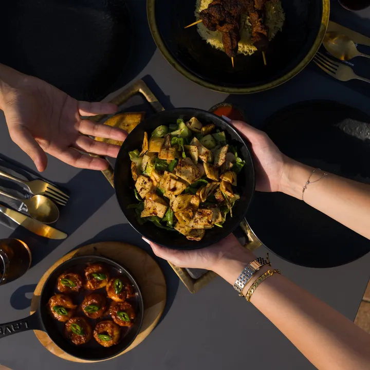 Hands passing a pan of pasta across a dinner table set with shared dishes